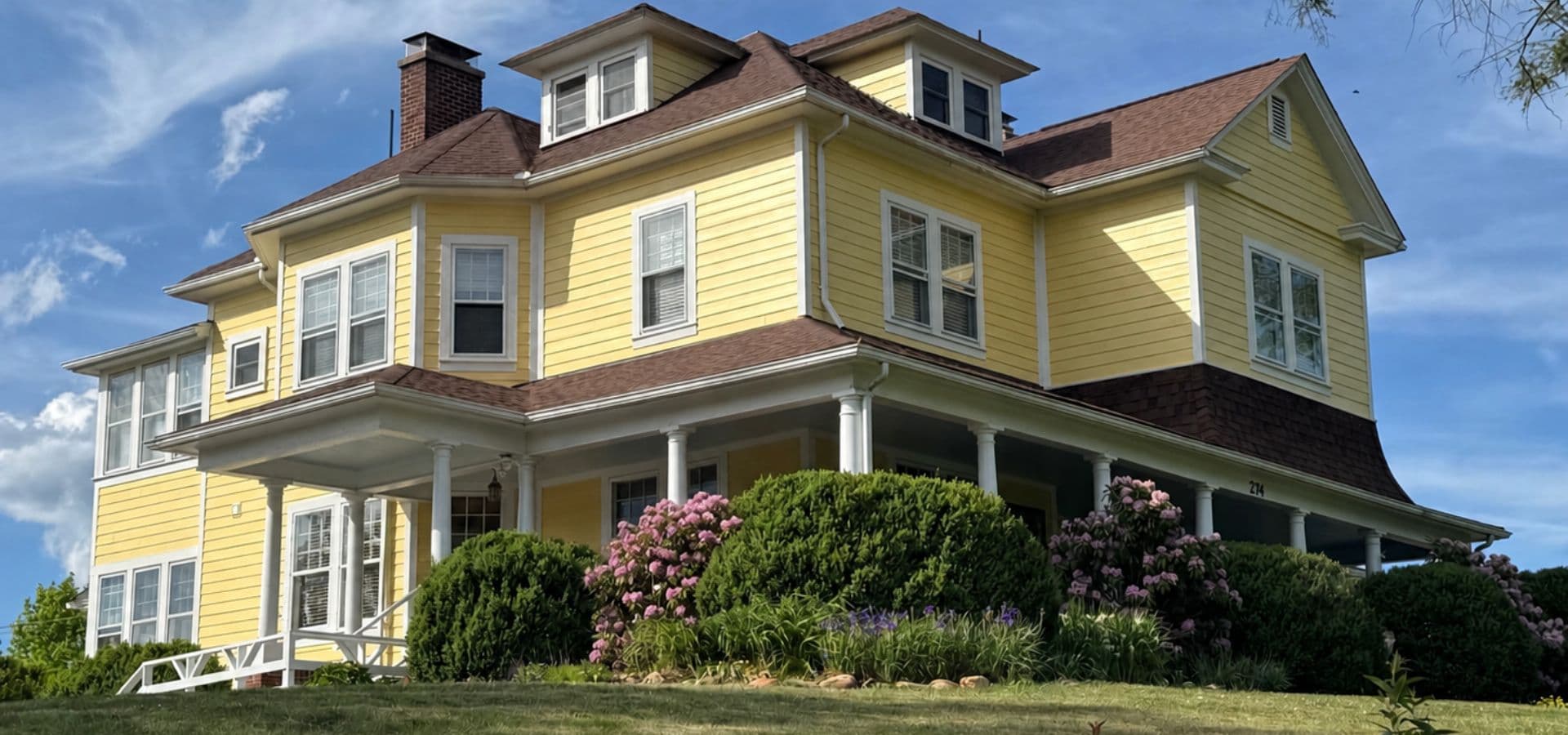 A large yellow Victorian house with a wraparound porch surrounded by green bushes and flowers.